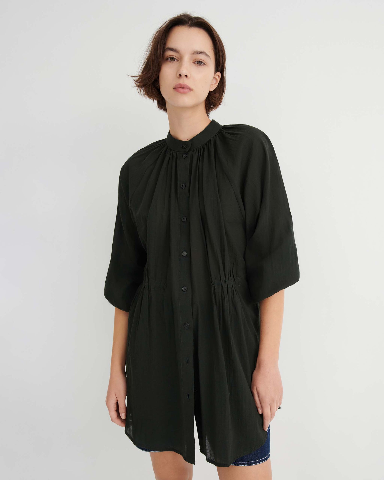 A person wearing a black, button-up, short-sleeved tunic with gathered detailing at the neckline and waist.