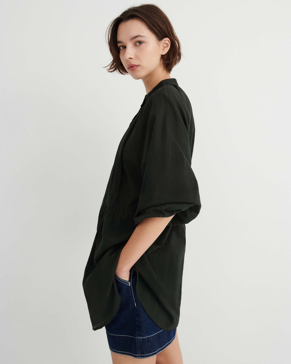 A person wearing an oversized dark green button-down shirt and blue denim shorts, standing in profile.