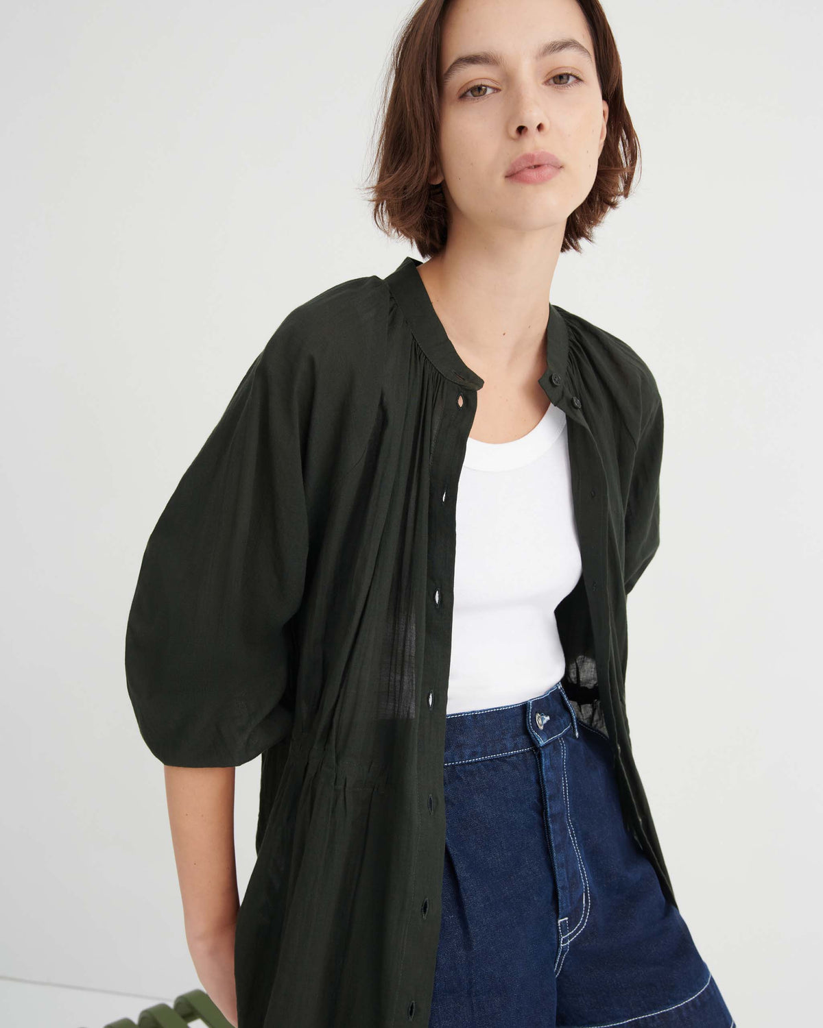 A person wearing a white tank top, dark blue jeans, and an unbuttoned dark green long-sleeved shirt.