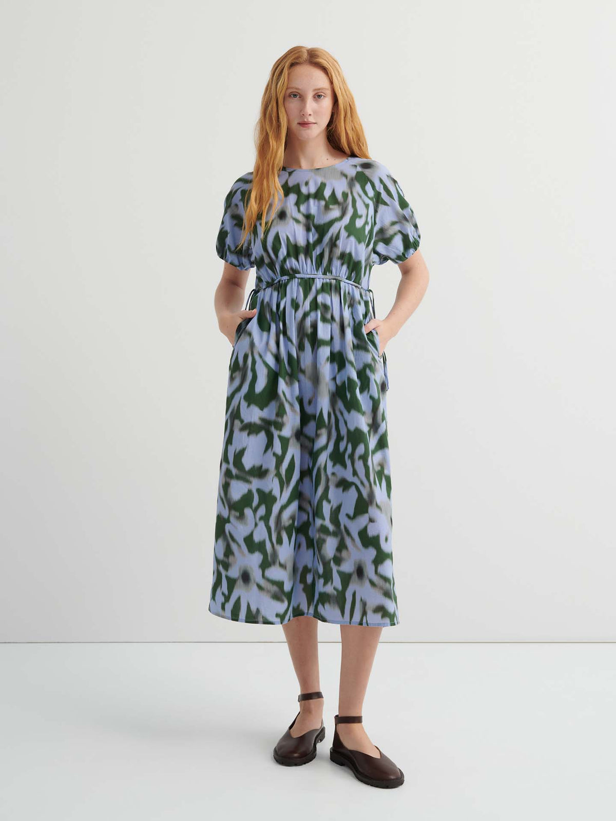 A woman stands against a plain background wearing the Illustrate Dress – Misty Floral, a loose-fitting, short-sleeve, green and blue patterned midi with pockets and dark flat shoes.