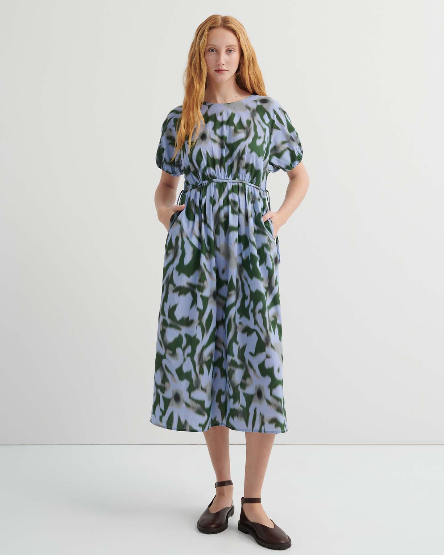 A woman stands against a plain background wearing the Illustrate Dress – Misty Floral, a loose-fitting, short-sleeve, green and blue patterned midi with pockets and dark flat shoes.