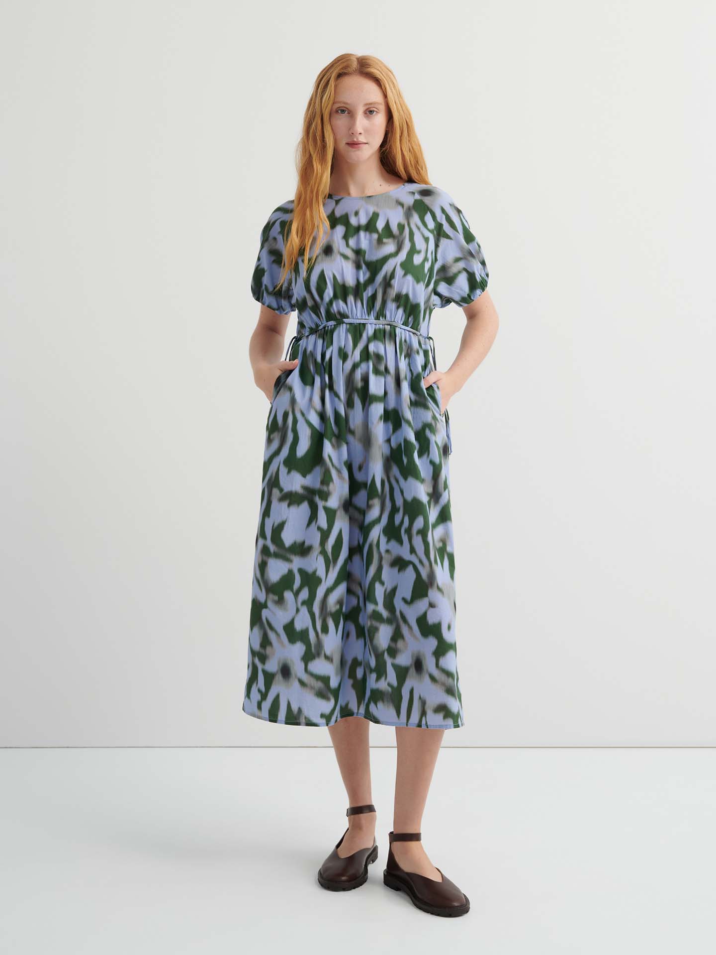 A woman stands against a plain background wearing the Illustrate Dress – Misty Floral, a loose-fitting, short-sleeve, green and blue patterned midi with pockets and dark flat shoes.