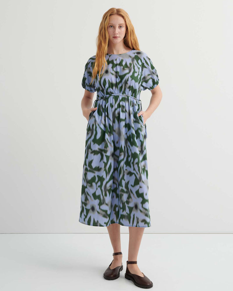 A woman stands against a plain background wearing the Illustrate Dress – Misty Floral, a loose-fitting, short-sleeve, green and blue patterned midi with pockets and dark flat shoes.