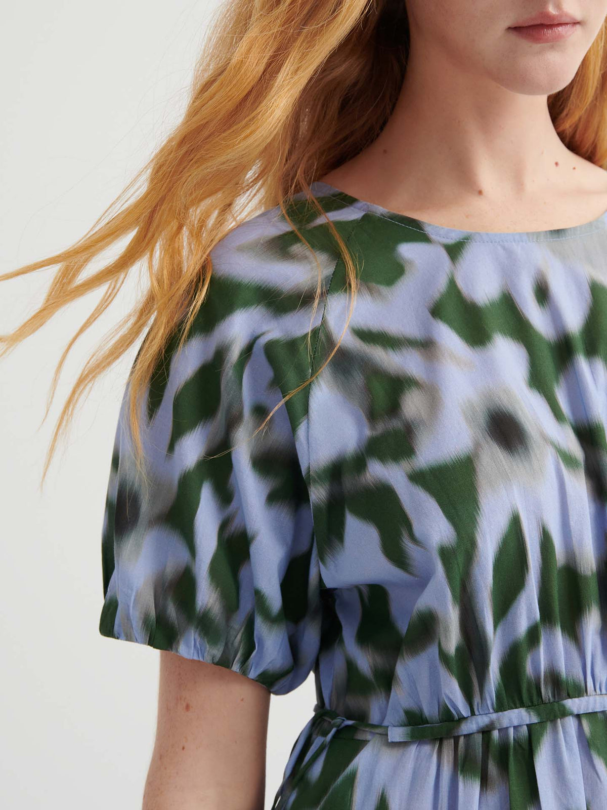 A woman with long, wavy red hair wears the Illustrate Dress – Misty Floral by Kowtow, a standard fit short-sleeved dress with a blue, green, and white abstract floral pattern. Only her upper body is shown.