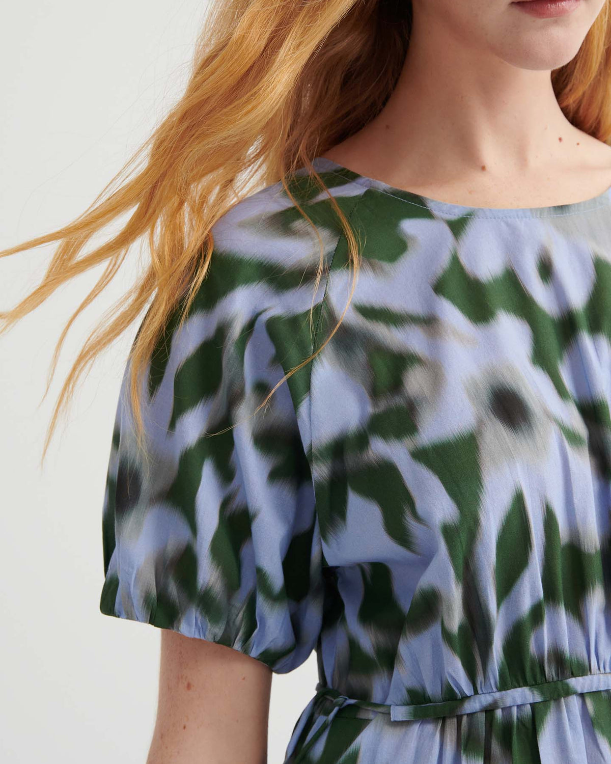 A woman with long, wavy red hair wears the Illustrate Dress – Misty Floral by Kowtow, a standard fit short-sleeved dress with a blue, green, and white abstract floral pattern. Only her upper body is shown.