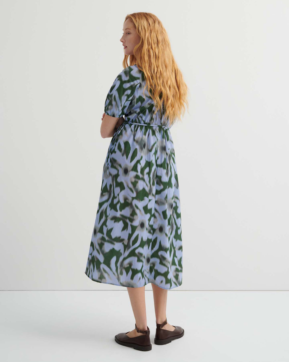 A woman with long red hair stands facing slightly away, wearing the Illustrate Dress – Misty Floral with a standard fit and brown shoes, set against a plain white background.