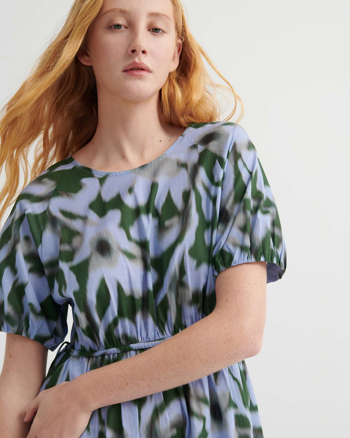 A woman with long red hair wears the Illustrate Dress – Misty Floral, which has a standard fit and a blue and green abstract floral pattern, standing against a plain light background.