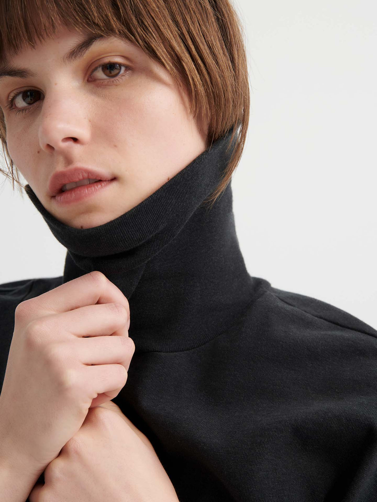 Person with short brown hair wears the Intent Top – Charcoal Marle in a relaxed fit, pulling up the collar with one hand while looking towards the camera.