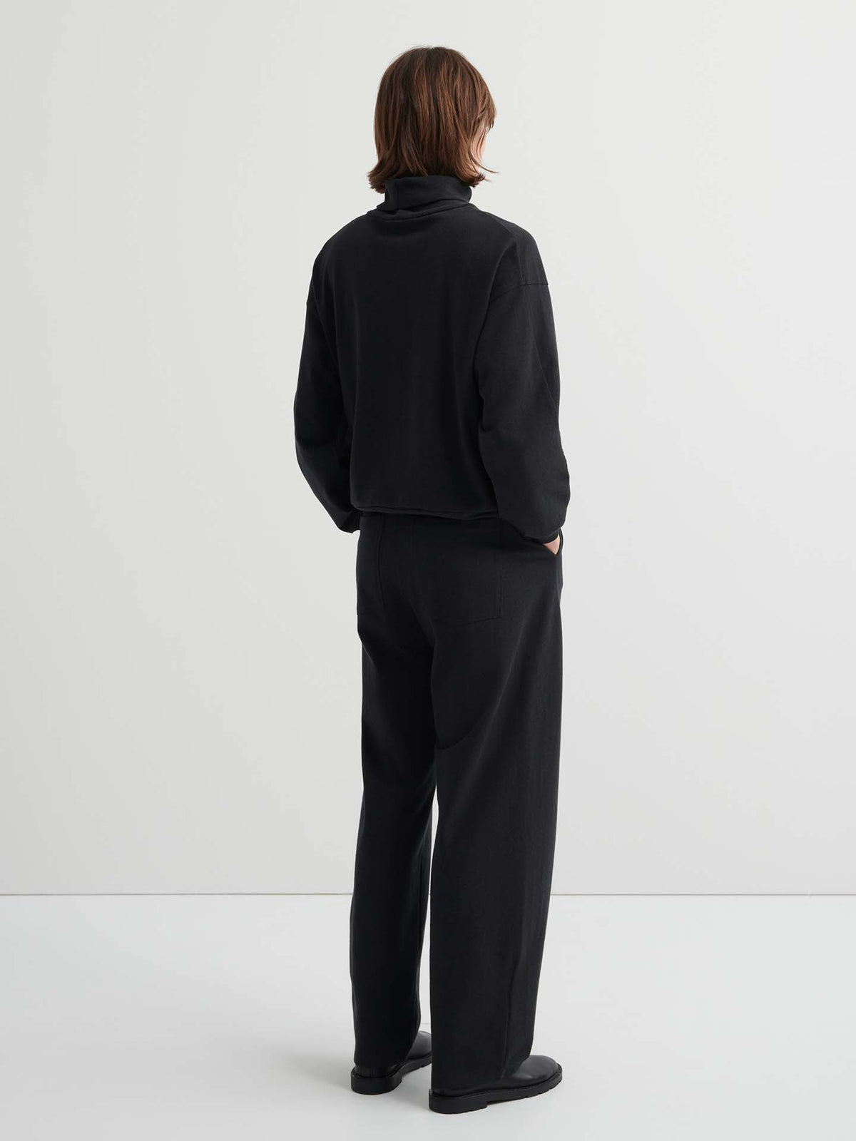 Person standing against a plain white background, wearing the Intent Top – Charcoal Marle with matching relaxed fit pants, facing away from the camera with hands in pockets. Check our size guide for the perfect fit.