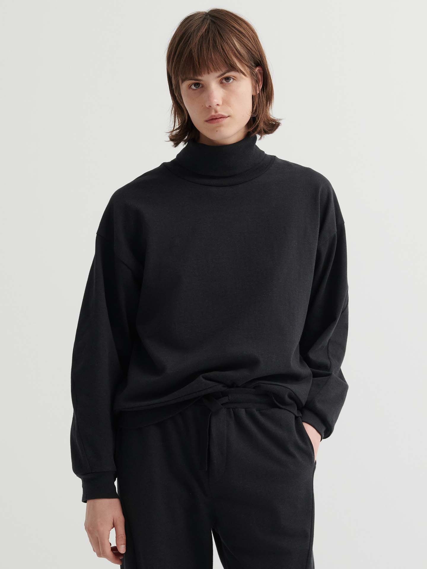 Person with straight brown hair wears a relaxed fit Intent Top in Charcoal Marle and matching black pants, standing against a plain light background. For the perfect fit, check our size guide.