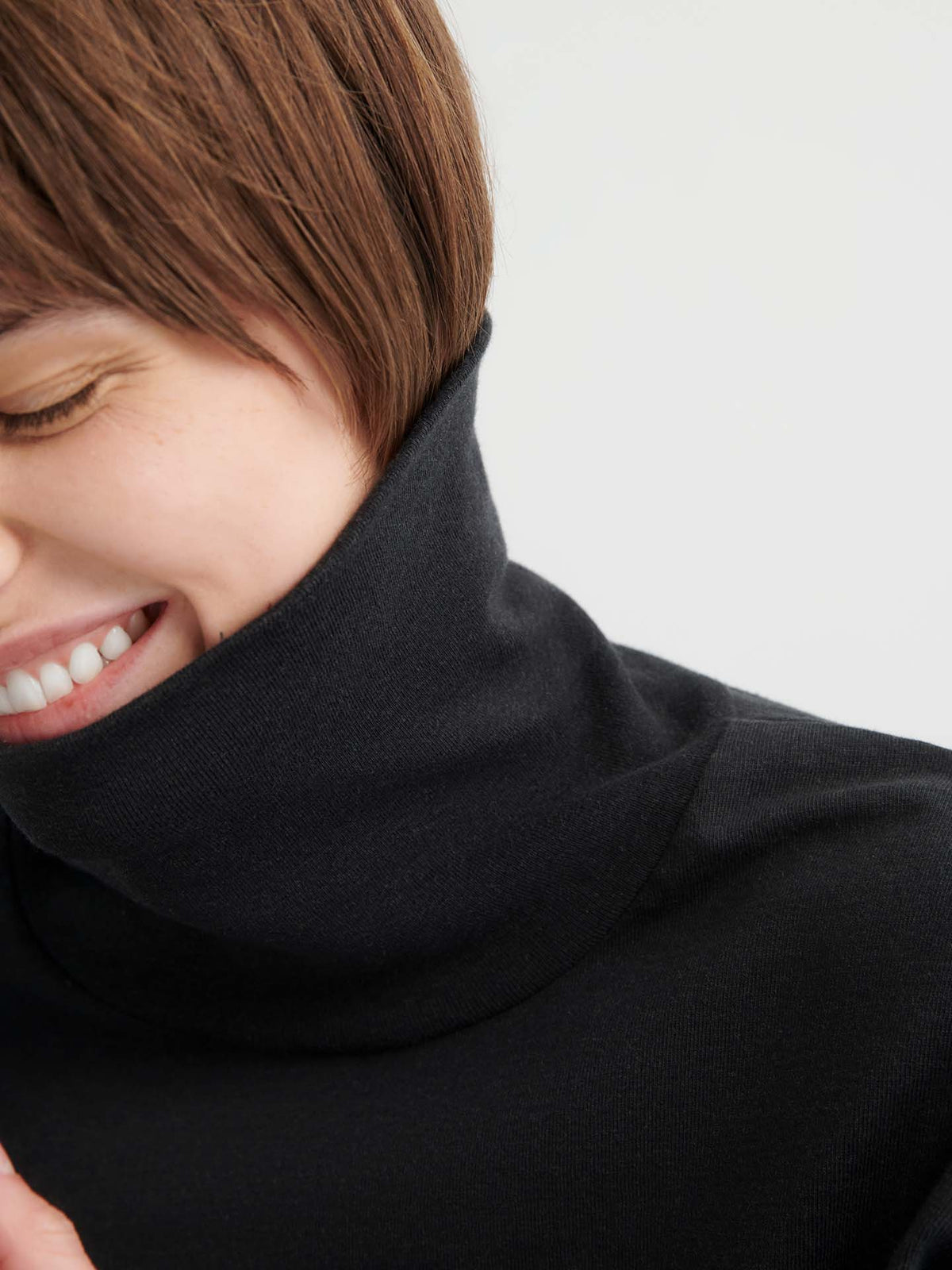 Close-up of a person with short brown hair wearing the relaxed fit Intent Top – Charcoal Marle turtleneck, smiling with eyes closed against a neutral background. Check our size guide to find your perfect fit.