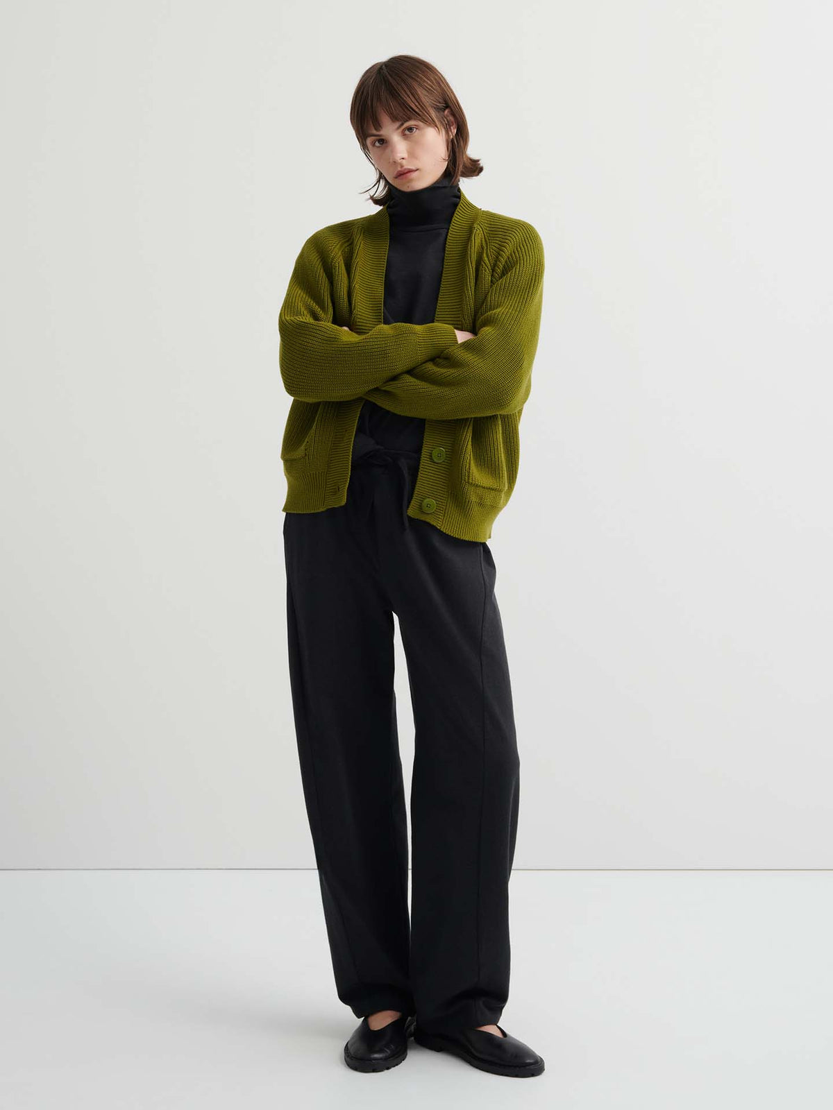 A person stands with arms crossed, wearing the relaxed fit Intent Top in Charcoal Marle over a black turtleneck and wide-leg black pants against a plain white background. See our size guide for your perfect fit.