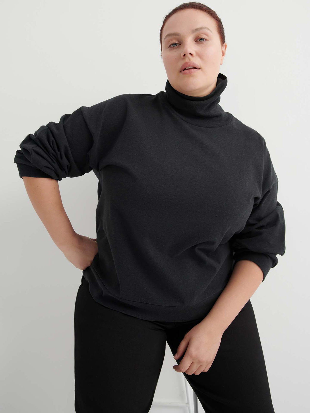 A person with light skin and dark hair pulled back wears the Intent Top – Charcoal Marle in a relaxed fit with black pants, standing against a plain light background, one hand on their hip. See size guide for your perfect fit.