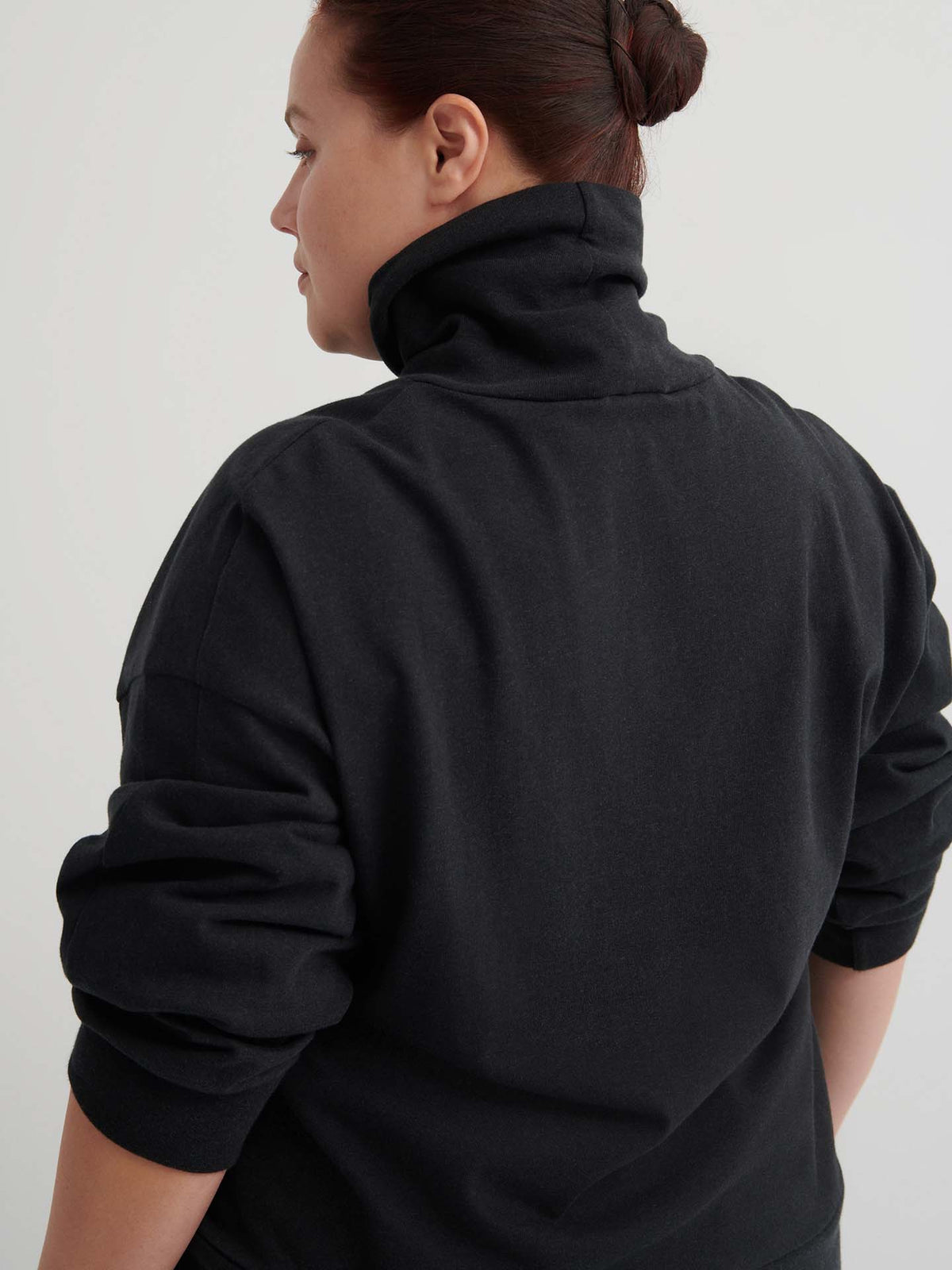 A person with dark hair in a bun, shown from the back and looking to the left, wears a relaxed-fit Intent Top – Charcoal Marle turtleneck sweatshirt.