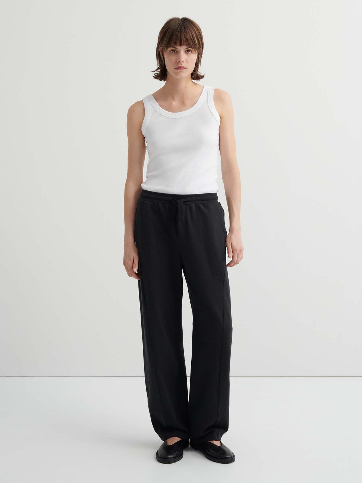 A person with short brown hair wears a white tank top, high-waist Intent Trackpant – Charcoal Marle in a relaxed fit, and black shoes, standing against a plain light background.