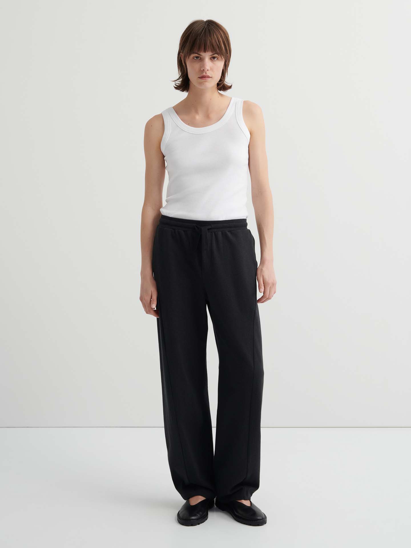 A person with short brown hair wears a white tank top, high-waist Intent Trackpant – Charcoal Marle in a relaxed fit, and black shoes, standing against a plain light background.