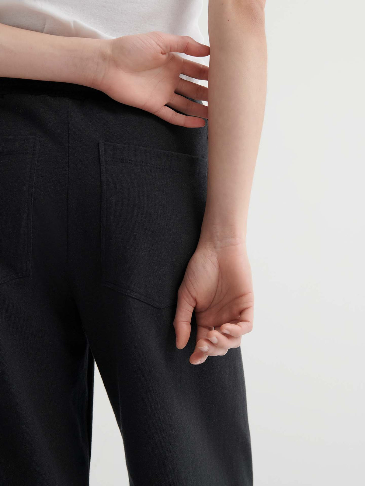 A person stands with their back to the camera, wearing Intent Trackpant – Charcoal Marle, paired with a white shirt. One arm is bent behind their back while the other hand rests on their lower back.