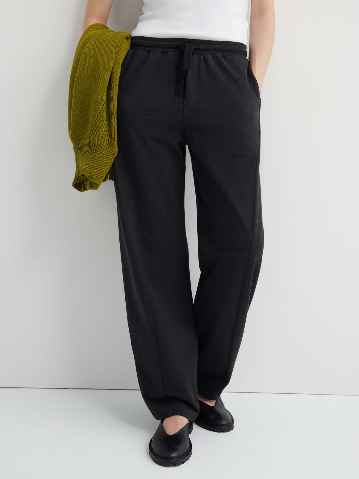 A person stands against a white background wearing the Intent Trackpant – Charcoal Marle, styled with a white top, black shoes, and a green sweater draped over one arm, showcasing effortless chic in this relaxed fit.