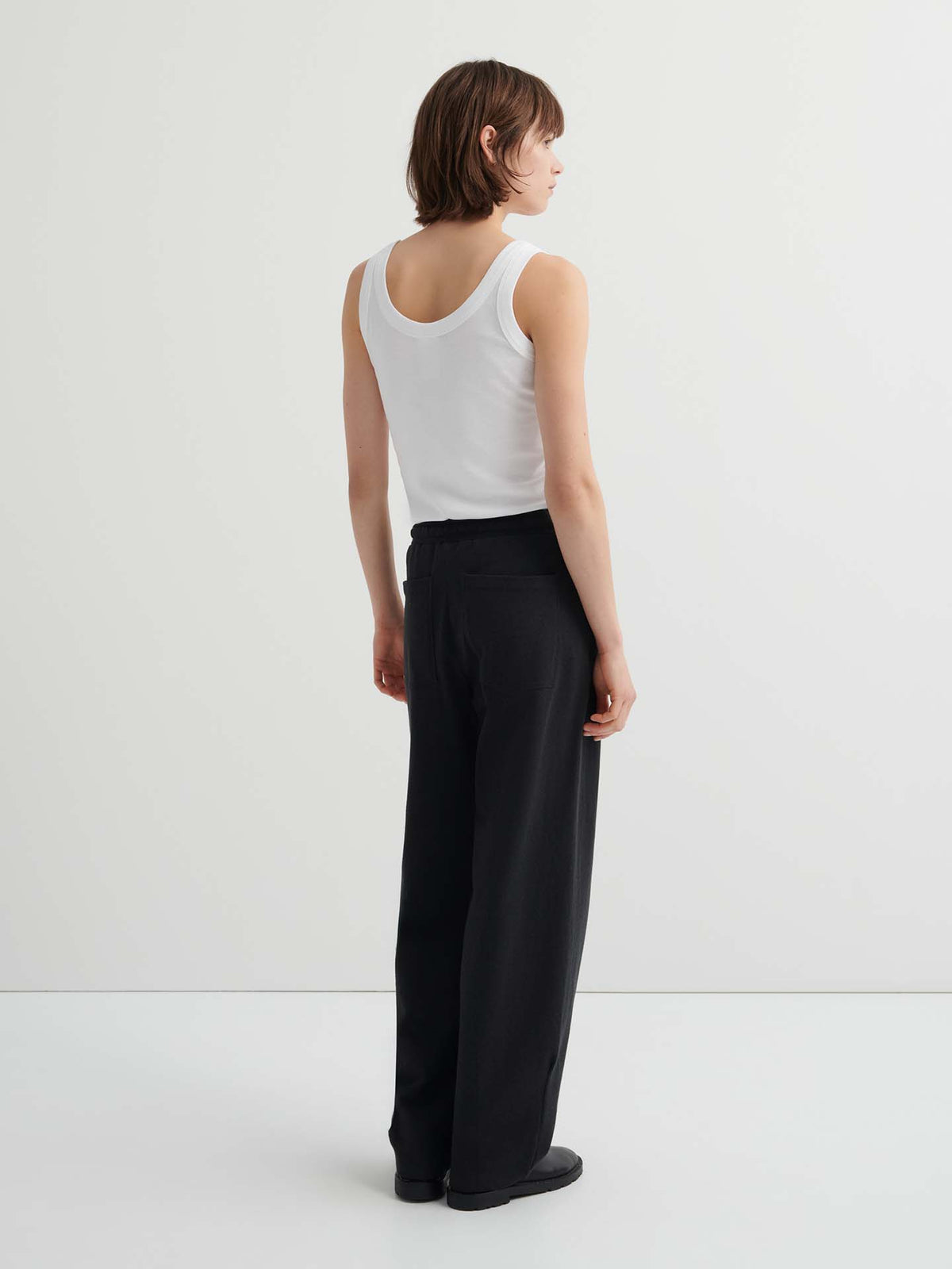 Person standing against a white background, facing away, in a white sleeveless top, Intent Trackpant – Charcoal Marle (high waist, relaxed fit), and black shoes.