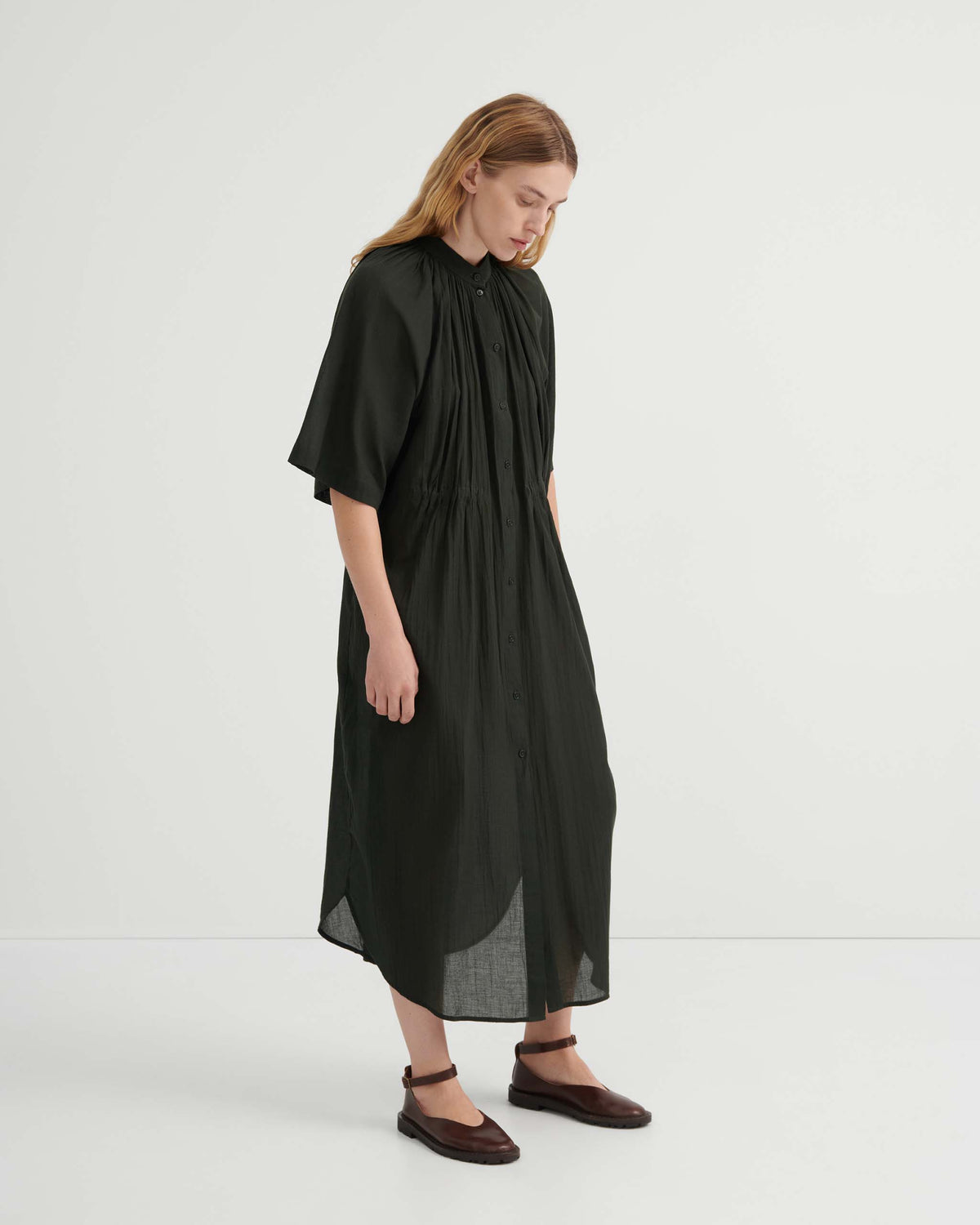 A woman wearing a button-front black midi dress with a gathered waist and dark brown ankle-strap shoes.