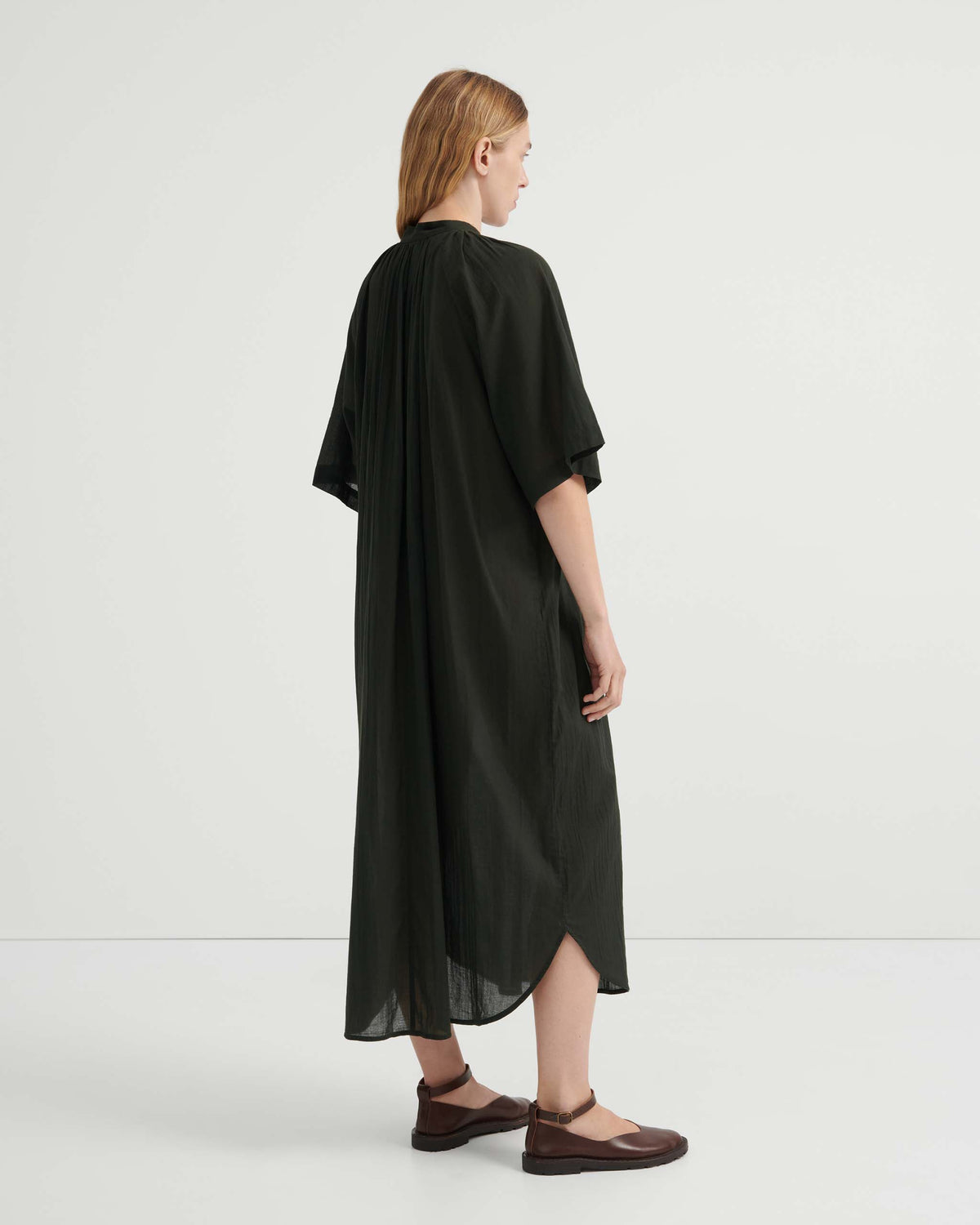 A person seen from behind wearing a loose, dark green midi dress with short sleeves and brown buckled flat shoes.