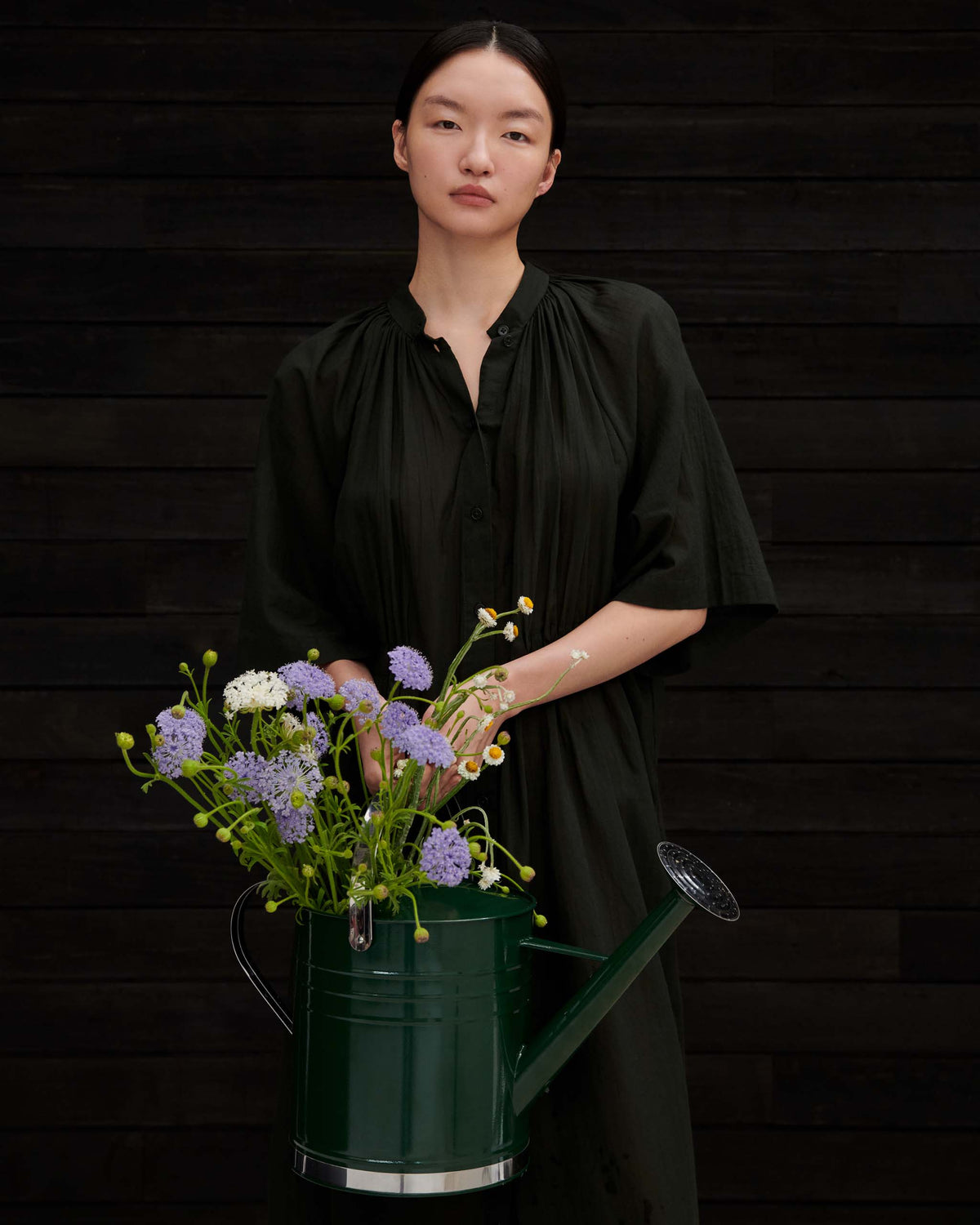 A person in a dark green dress holding a green watering can filled with blue and white flowers.