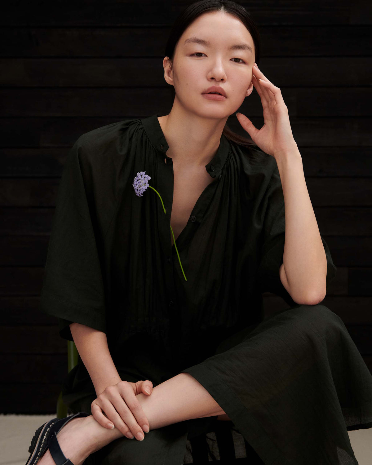 A person wearing a dark green, button-down midi dress with a single purple flower tucked into the neckline.