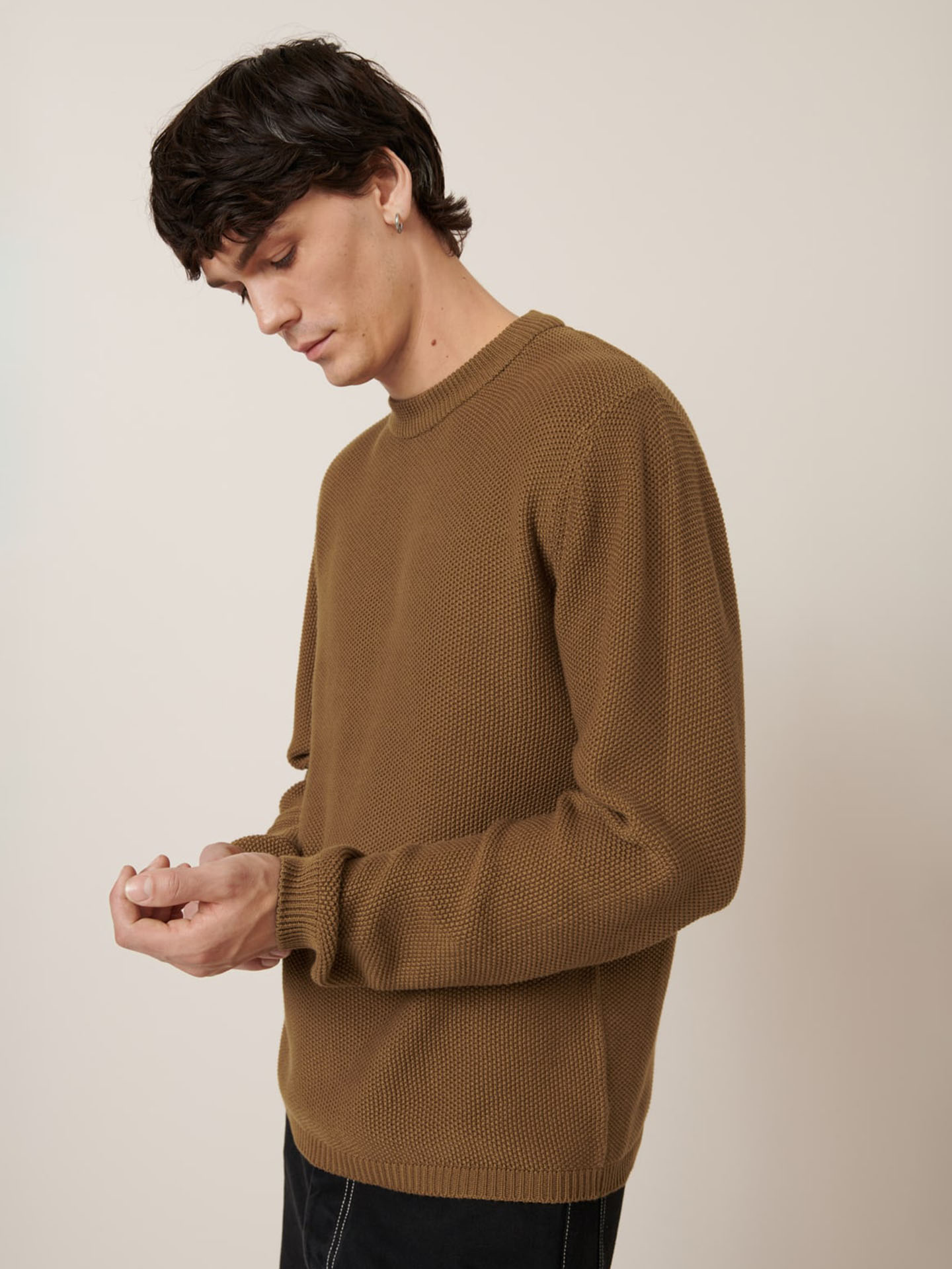 A person with short dark hair wears the Kowtow Judd Crew in Dark Spice, a brown textured sweater with a relaxed fit, and looks down while adjusting their sleeve against a plain beige background.