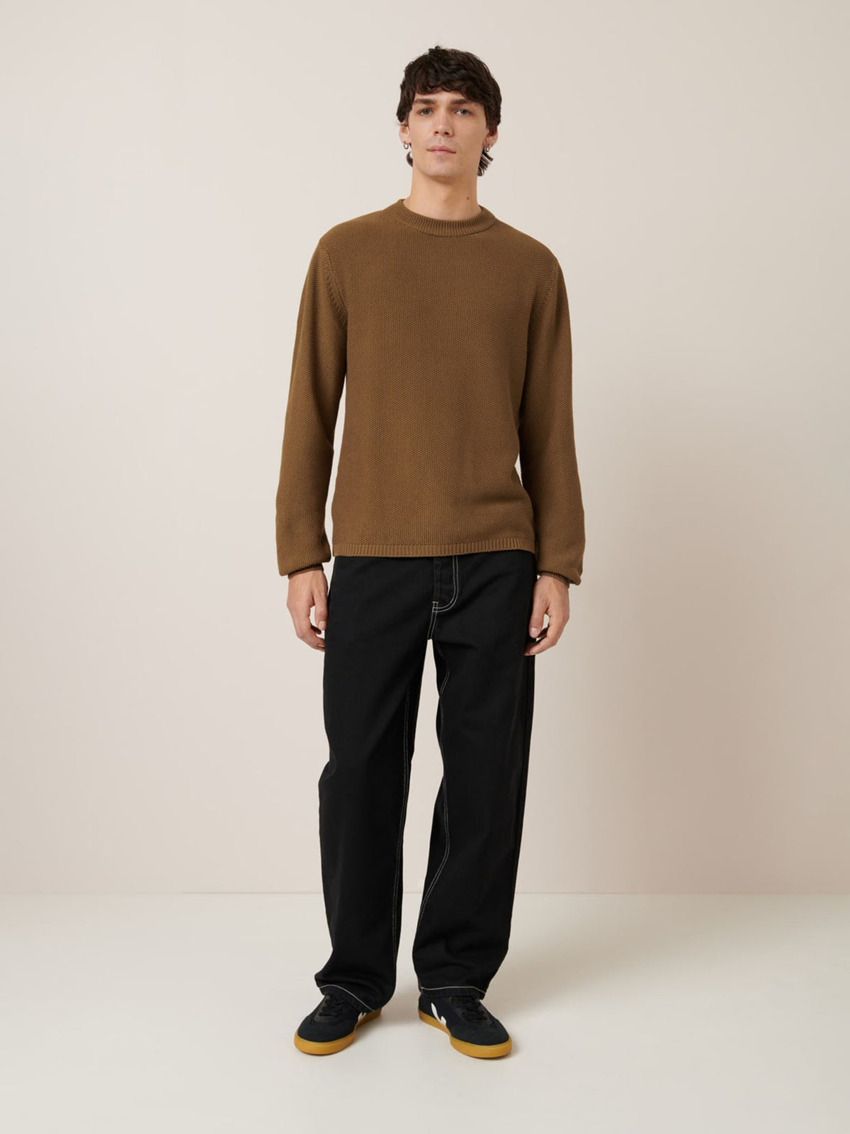 A young man stands against a plain, light background wearing the Kowtow Judd Crew in Dark Spice, styled in a relaxed fit with black pants and black sneakers featuring gum soles.