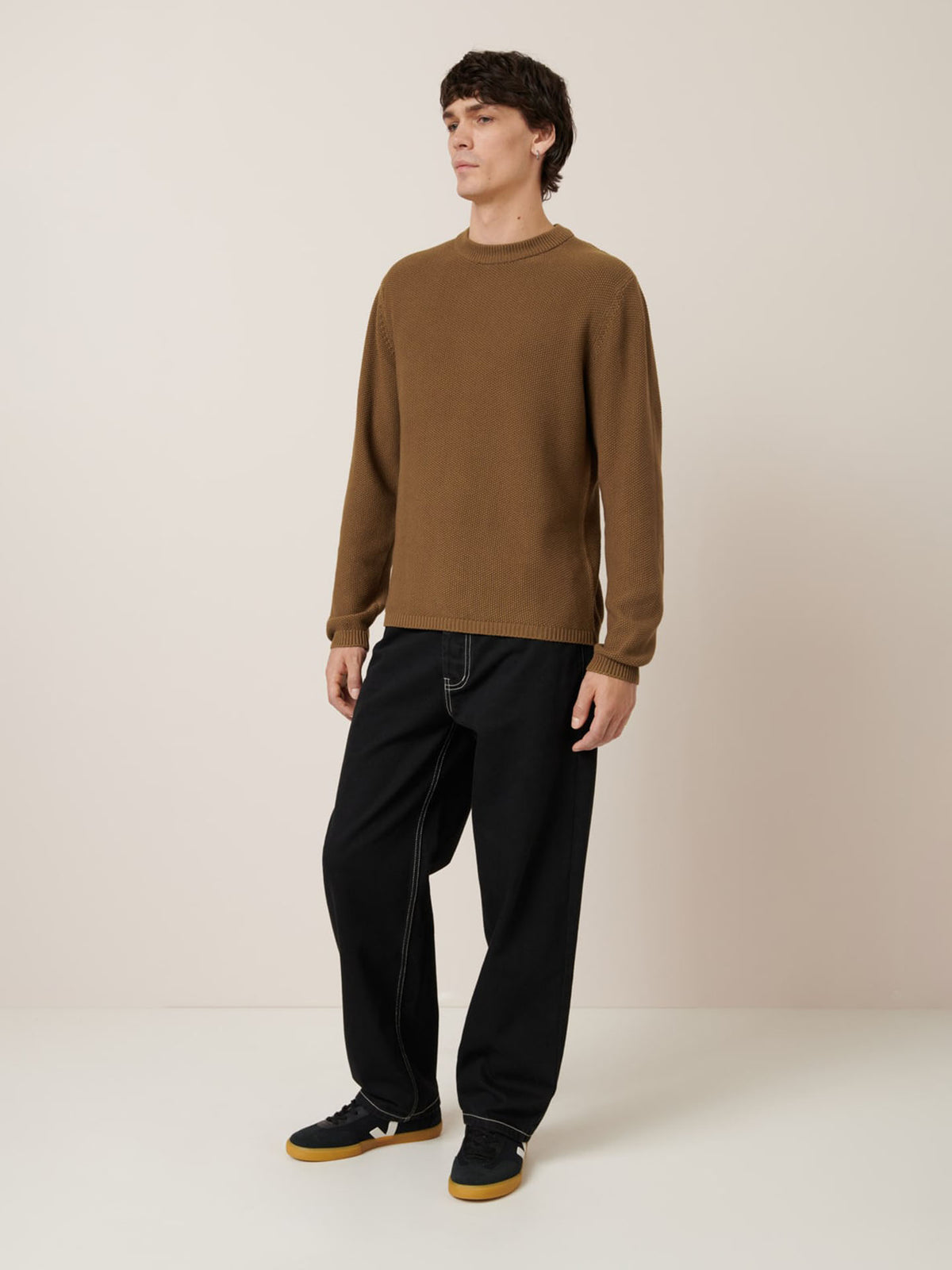A man stands against a plain background wearing the Kowtow Judd Crew in Dark Spice, relaxed fit black pants with white stitching highlighting the pattern, and black sneakers with gum soles.