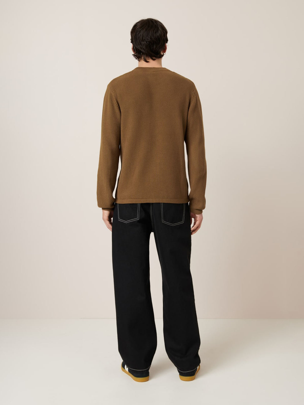 A person faces away from the camera, wearing the Kowtow Judd Crew in Dark Spice and black wide-leg pants with white stitching, all in a relaxed fit against a plain light background.