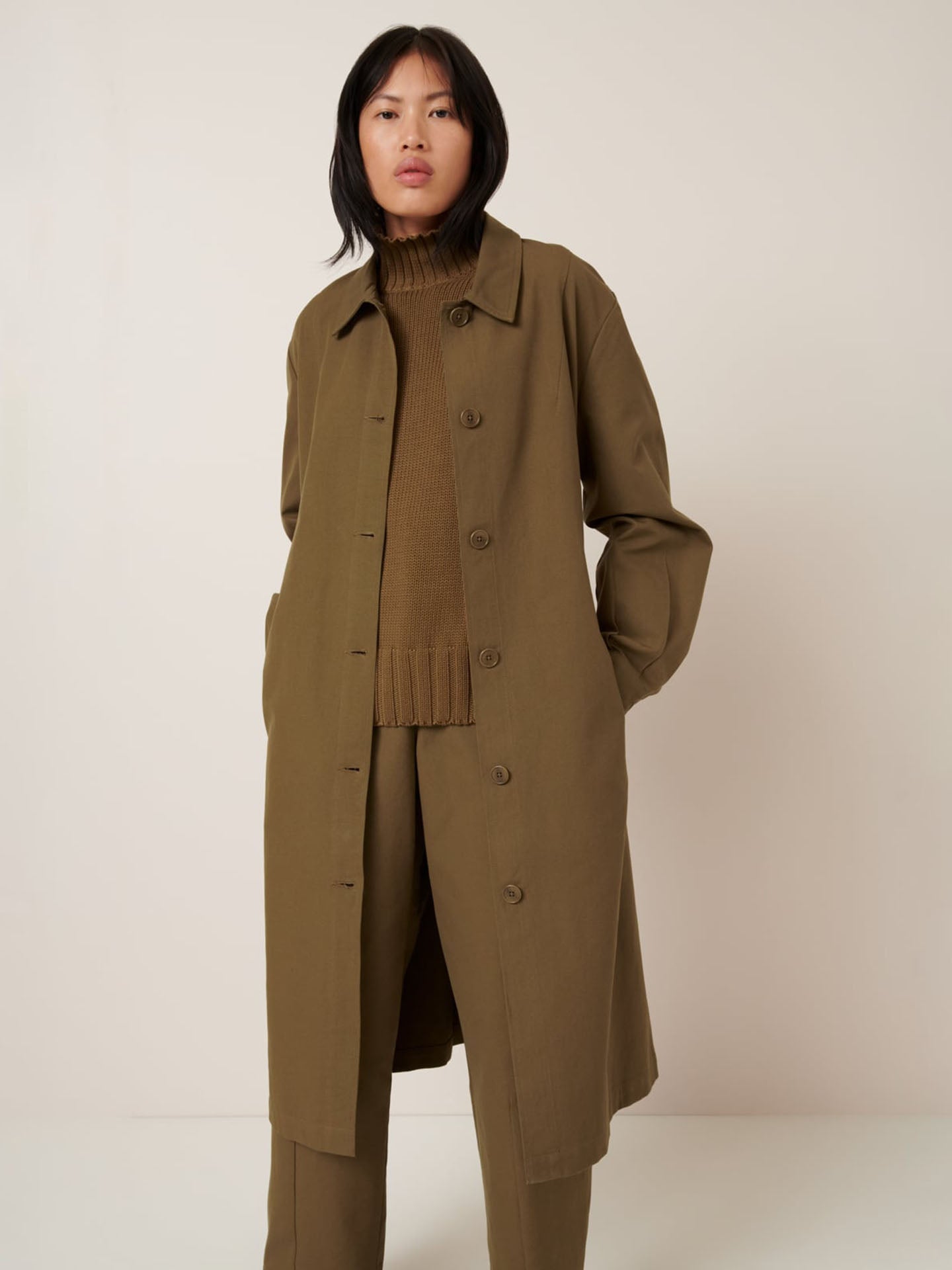 A person wears the Kowtow Landscape Trench in Dark Spice, an oversized olive green coat, over a brown turtleneck and matching trousers, standing against a plain light background.
