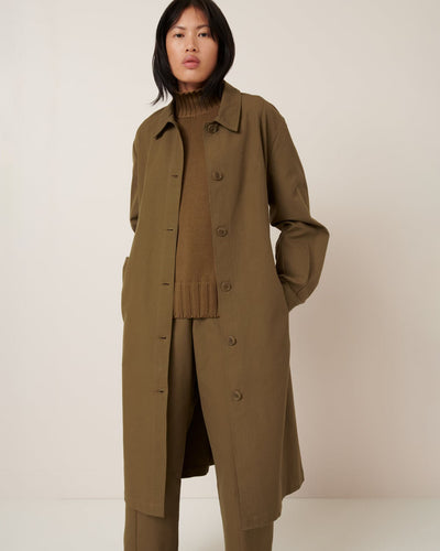 A person wears the Kowtow Landscape Trench in Dark Spice, an oversized olive green coat, over a brown turtleneck and matching trousers, standing against a plain light background.