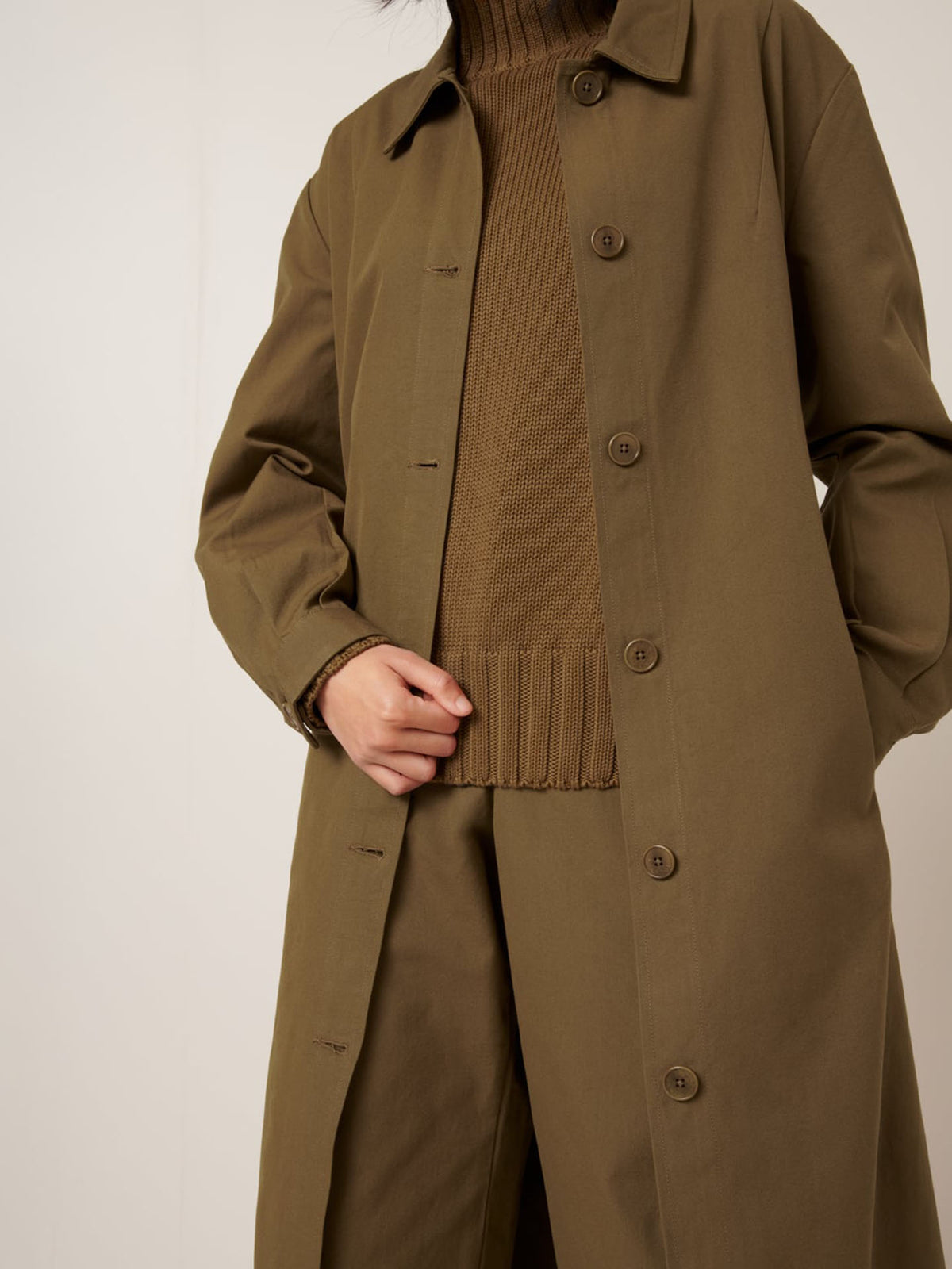 A person wearing the Kowtow Landscape Trench in Dark Spice with a matching sweater and pants, holding the coat lapel; their face is not visible.