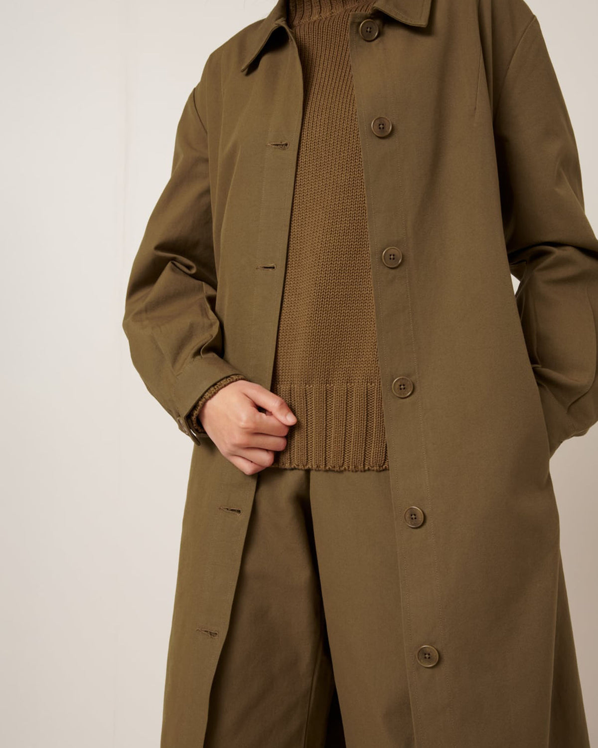 A person wearing the Kowtow Landscape Trench in Dark Spice with a matching sweater and pants, holding the coat lapel; their face is not visible.