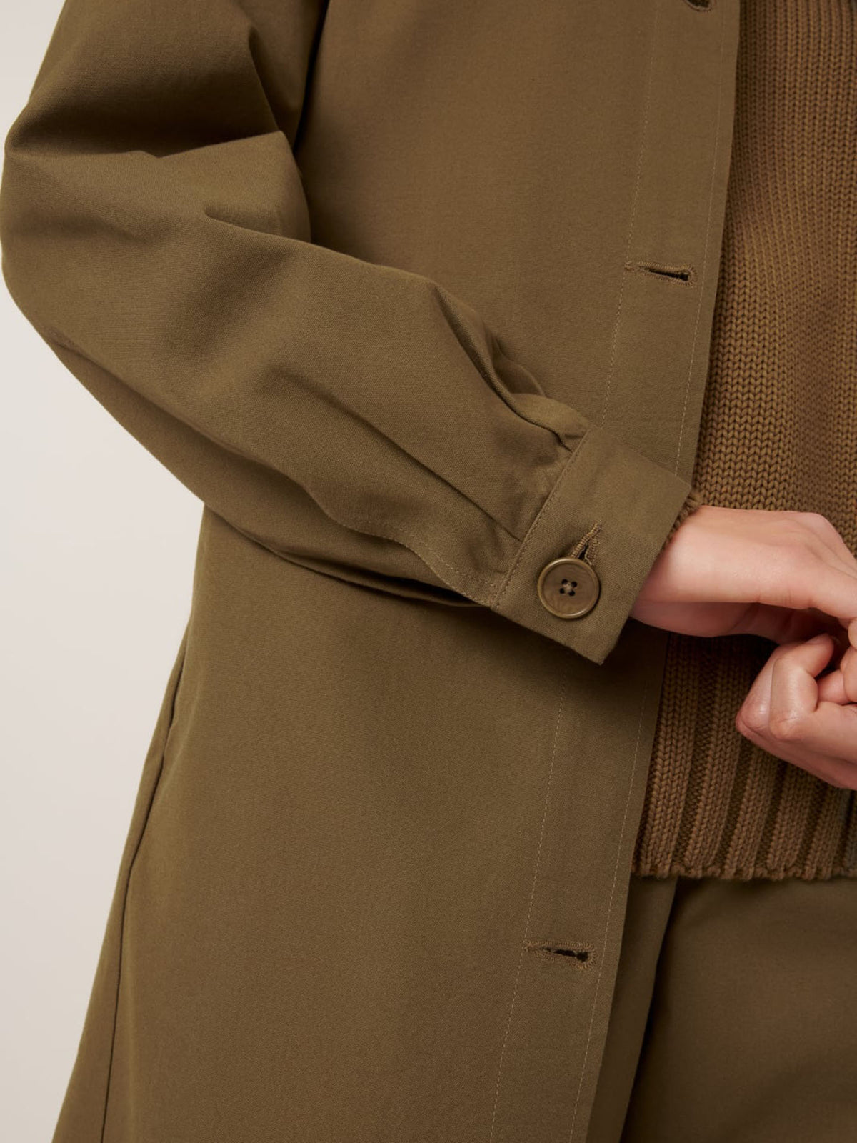 Close-up of a person wearing the Kowtow Landscape Trench in Dark Spice, an oversized buttoned coat, layered over a brown knit sweater with their hand partially visible and fingers intertwined.