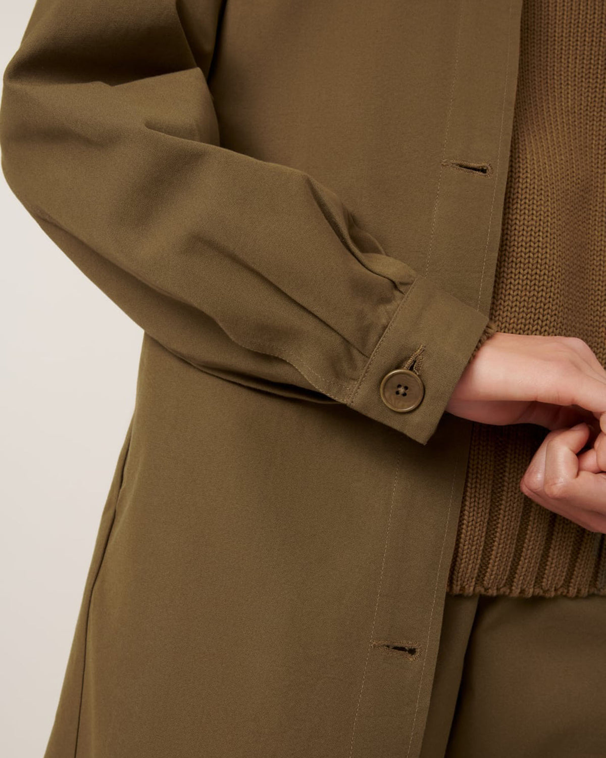 Close-up of a person wearing the Kowtow Landscape Trench in Dark Spice, an oversized buttoned coat, layered over a brown knit sweater with their hand partially visible and fingers intertwined.