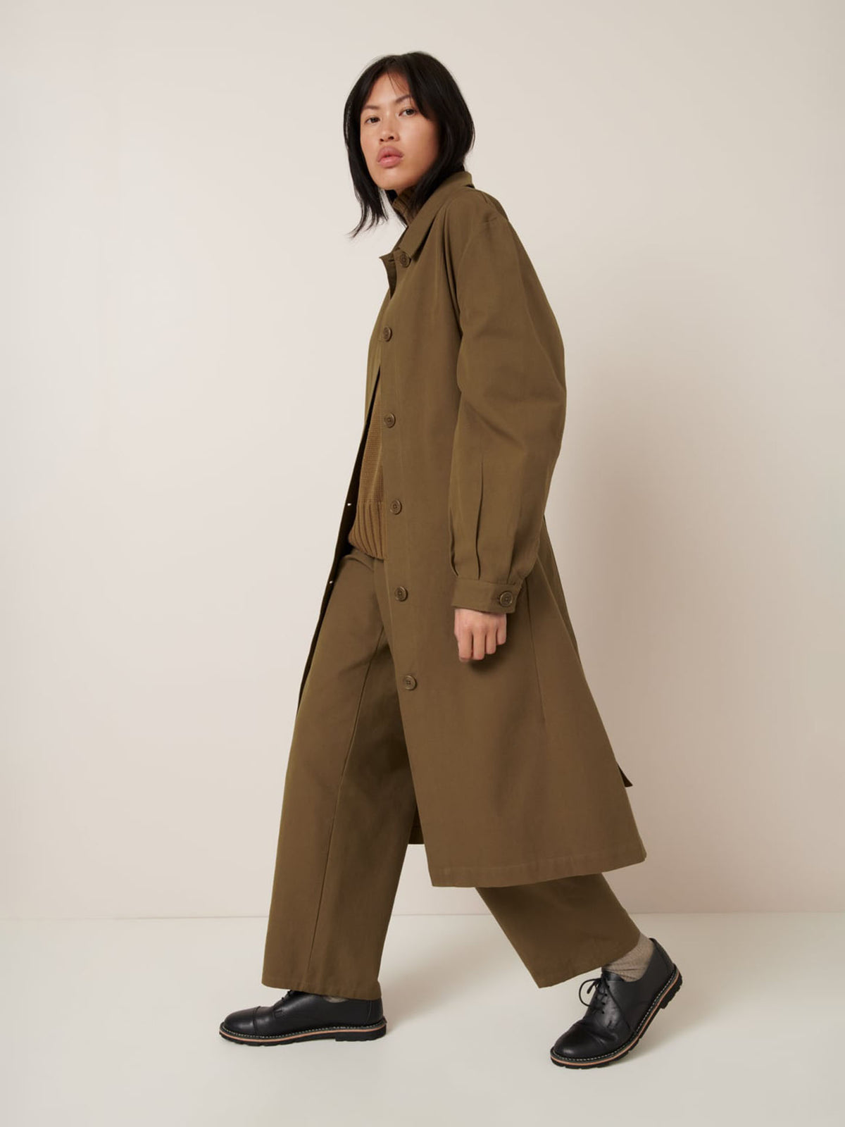 A person wears the Kowtow Landscape Trench in Dark Spice, styled in an oversized fit with wide-leg pants and black shoes, standing against a plain off-white background.