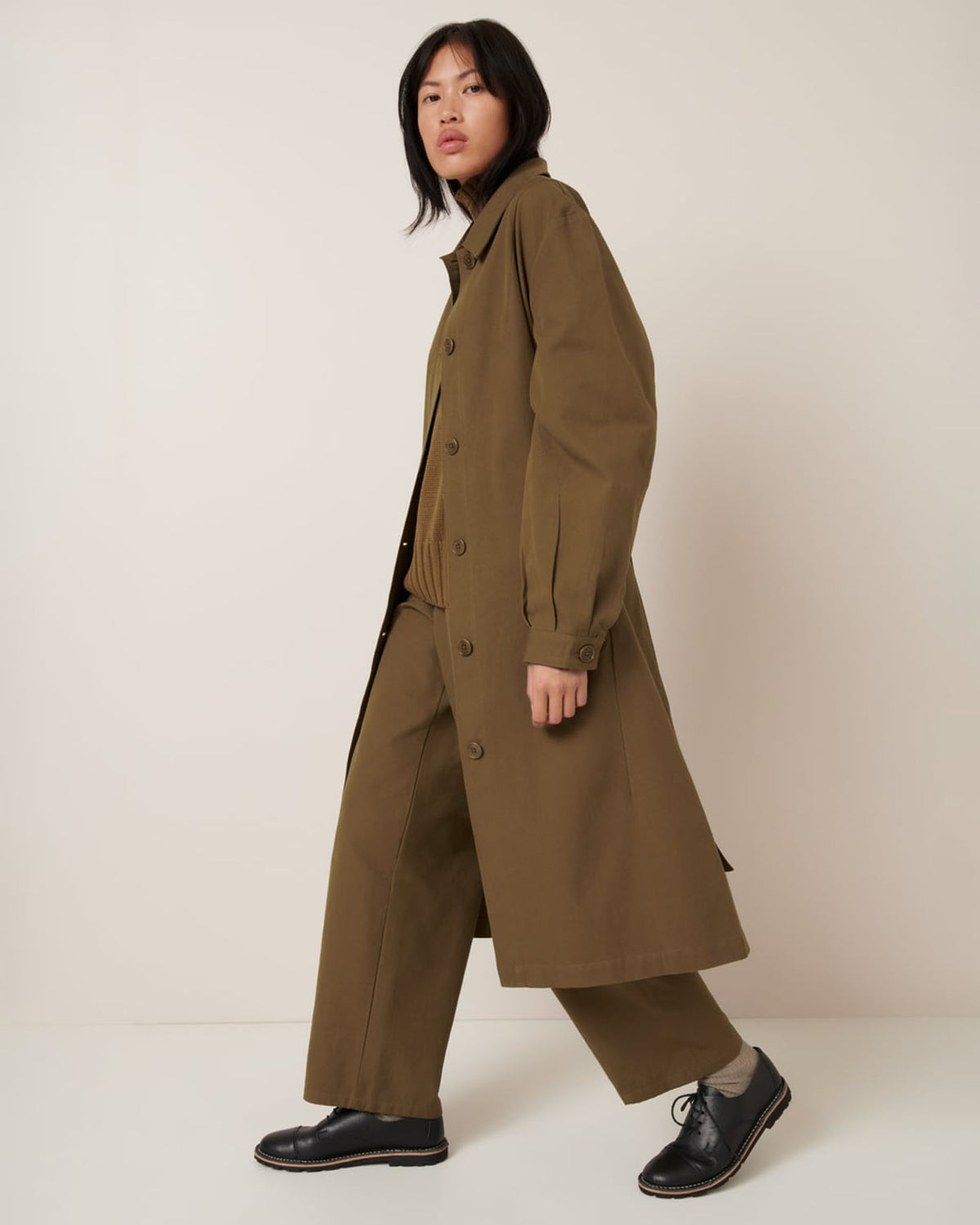 A person wears the Kowtow Landscape Trench in Dark Spice, styled in an oversized fit with wide-leg pants and black shoes, standing against a plain off-white background.