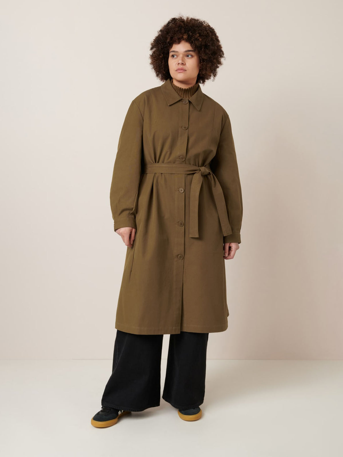 A person stands against a plain background wearing the Landscape Trench in Dark Spice by Kowtow, styled in an oversized fit and belted at the waist, paired with black wide-leg pants and black shoes with tan soles.