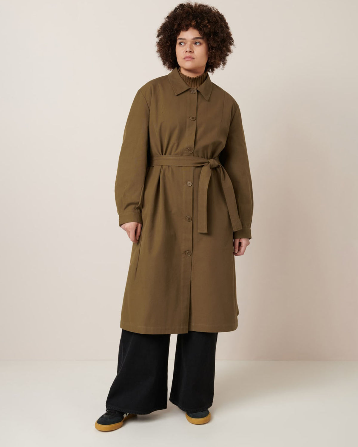A person stands against a plain background wearing the Landscape Trench in Dark Spice by Kowtow, styled in an oversized fit and belted at the waist, paired with black wide-leg pants and black shoes with tan soles.