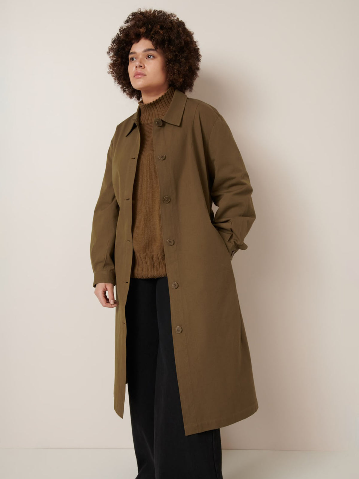 A person stands against a plain light wall wearing Kowtow’s Landscape Trench in Dark Spice, styled with a brown sweater and black pants.