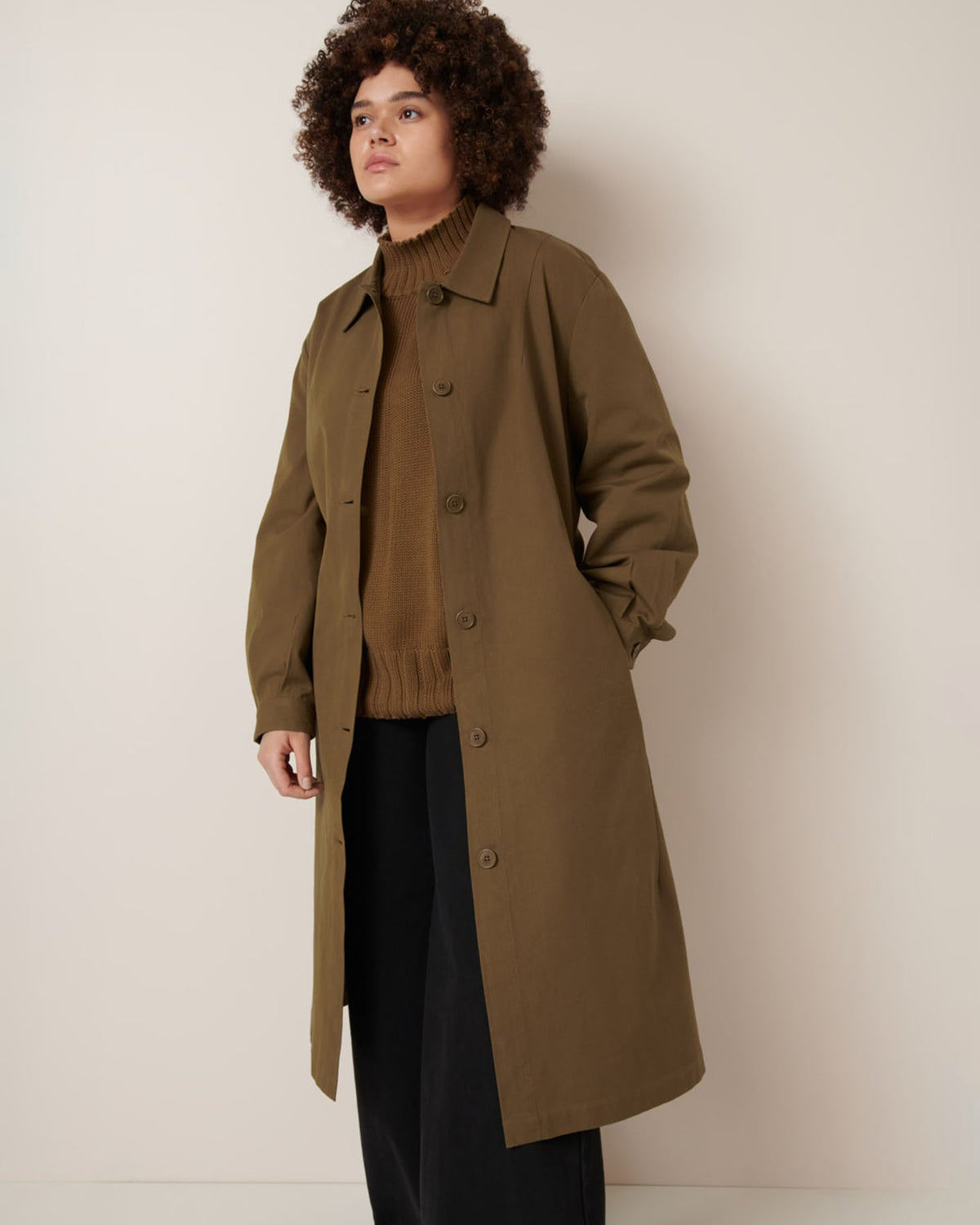 A person stands against a plain light wall wearing Kowtow’s Landscape Trench in Dark Spice, styled with a brown sweater and black pants.