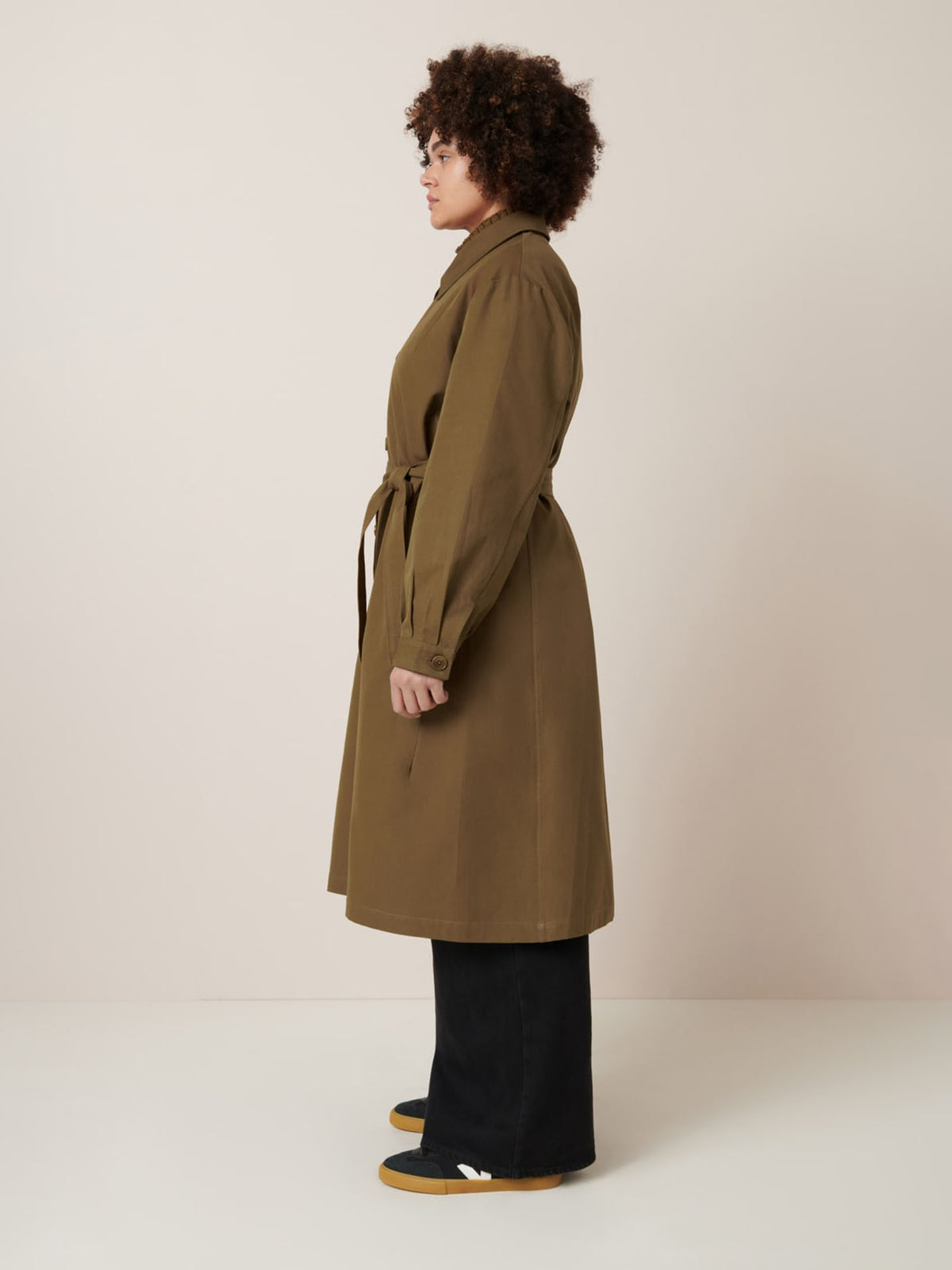 A person stands in profile wearing Kowtow&#39;s Landscape Trench in Dark Spice, styled with black pants and yellow shoes against a neutral background.