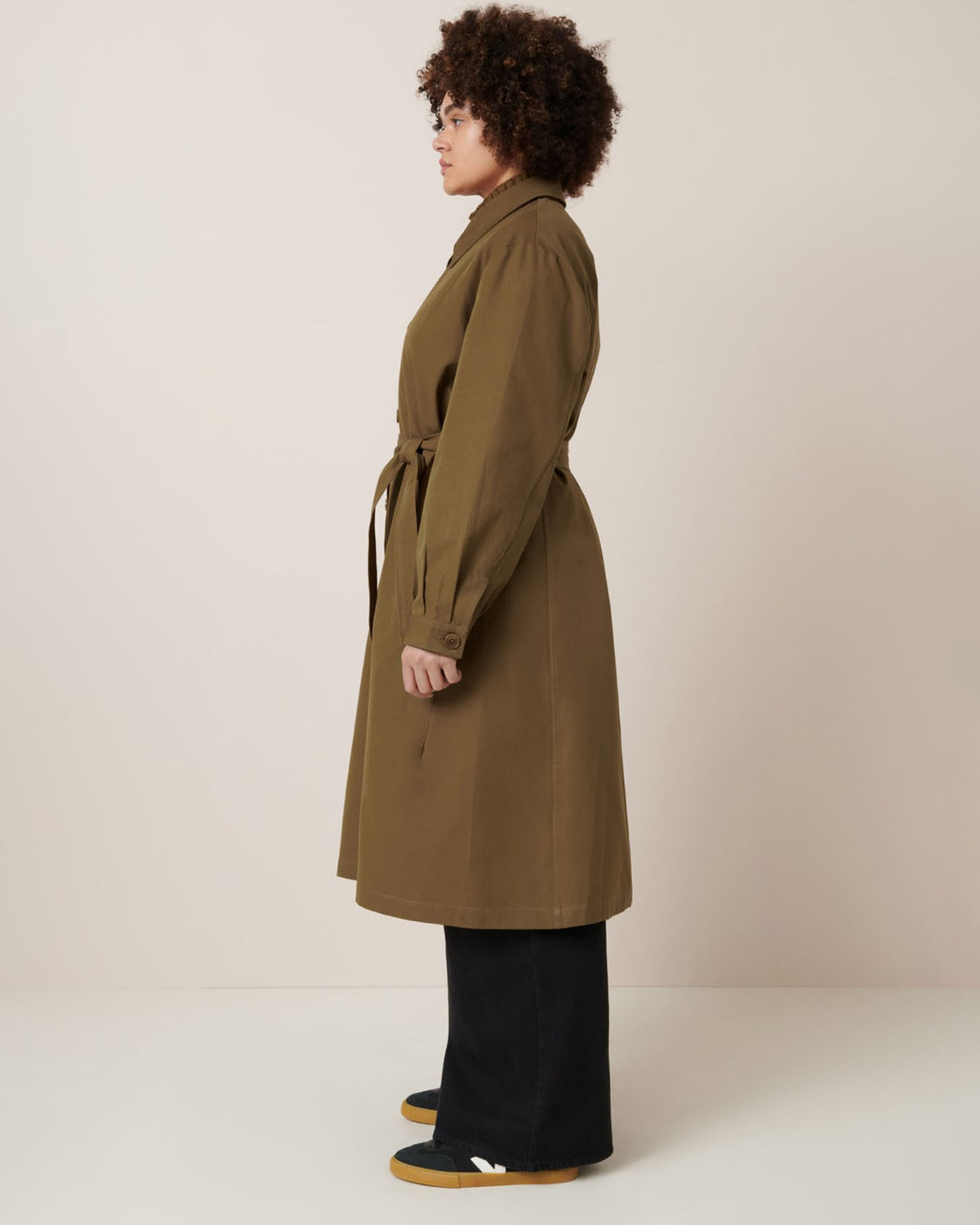 A person stands in profile wearing Kowtow's Landscape Trench in Dark Spice, styled with black pants and yellow shoes against a neutral background.
