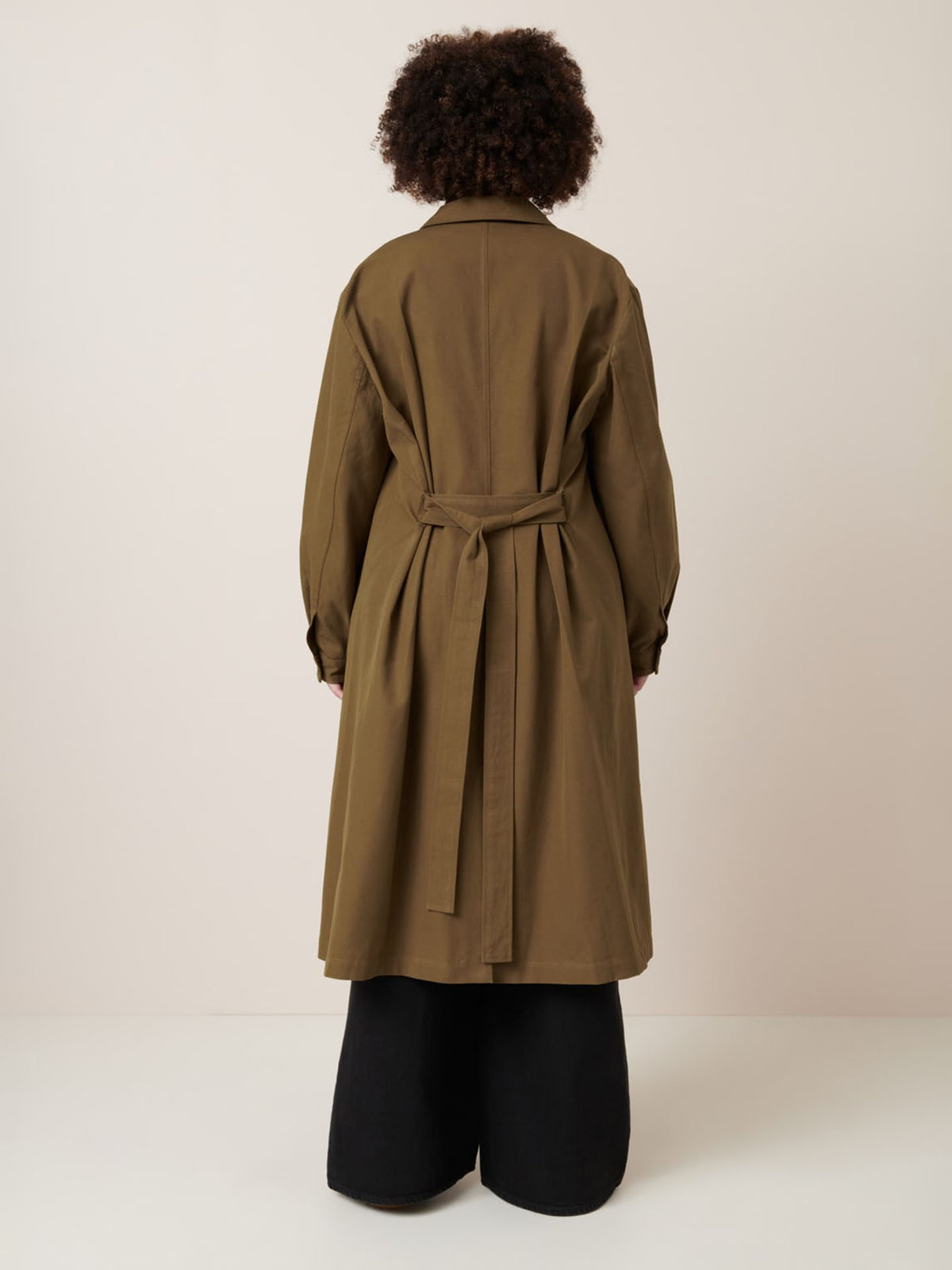 Facing away, a person wears the Kowtow Landscape Trench in Dark Spice with an oversized fit over wide-leg black pants, set against a plain light background.