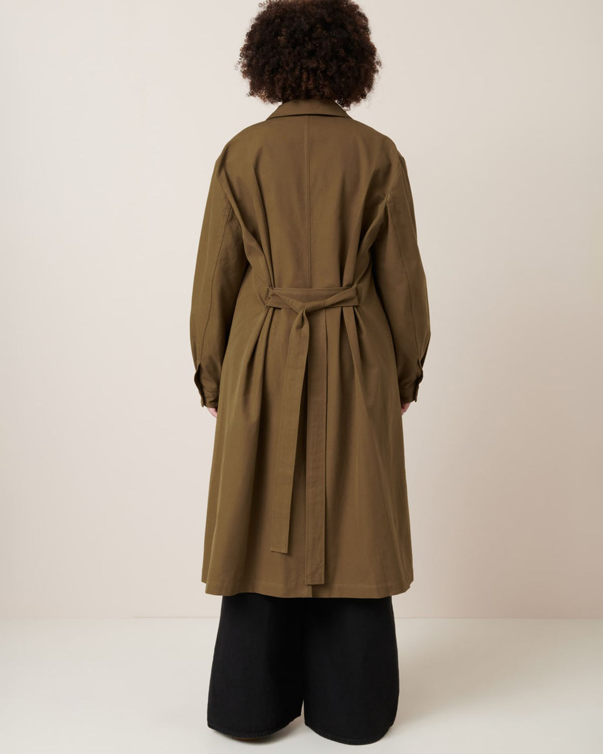 Facing away, a person wears the Kowtow Landscape Trench in Dark Spice with an oversized fit over wide-leg black pants, set against a plain light background.