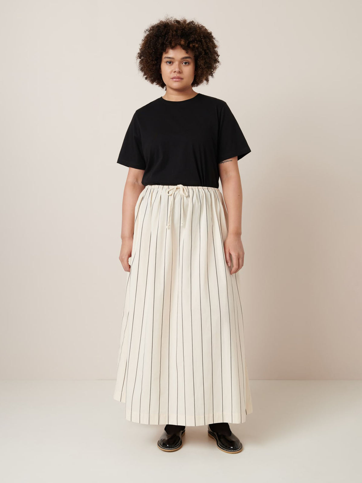 A person showcases a relaxed fit in a black T-shirt paired with the Kowtow Linear Skirt in Greige Pinstripe, against a plain background. The ensemble reflects precision in garment measurements for an effortlessly chic style, perfect for any occasion.