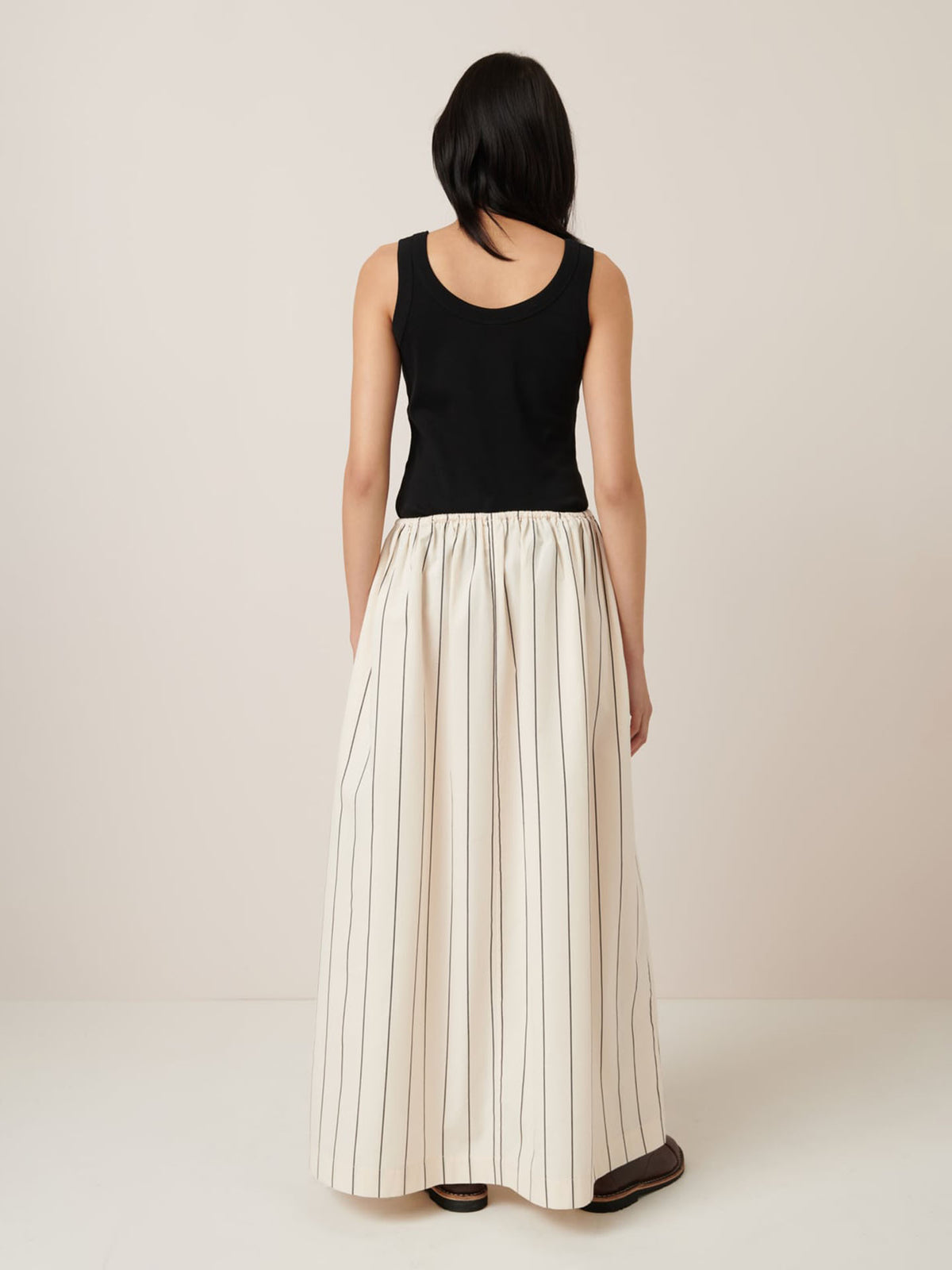 A person with long dark hair, wearing a black sleeveless top and the Kowtow Linear Skirt in greige pinstripe, stands facing away in a neutral setting.