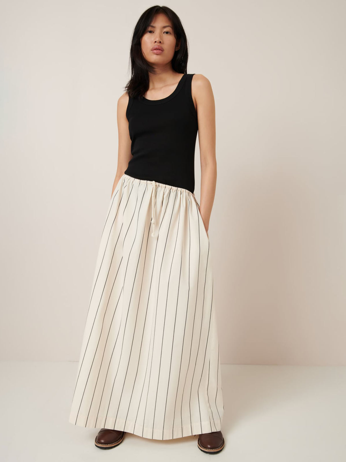 A model showcases the Linear Skirt – Greige Pinstripe by Kowtow, wearing a black tank top with the elegantly relaxed skirt. Its vertical stripes and hands-in-pocket design stand out against a plain background.